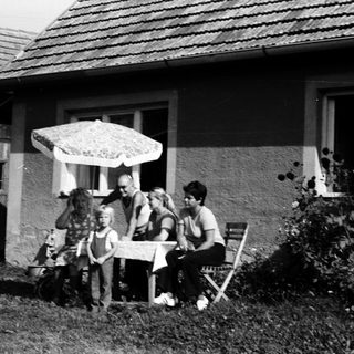 Family sitting outdoors near a house with umbrella.