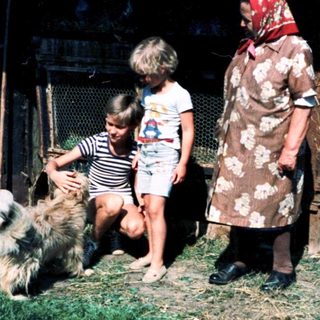 Rural scene: woman, children petting dog near farm structures.