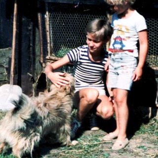 Rural scene: woman, children petting dog near farm structures.