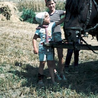 Rural scene with child and adult posing beside hay bale and harnessed horse.