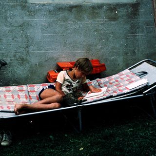 Child sitting on cot in makeshift indoor space.