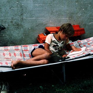 Child sitting on cot in makeshift indoor space.