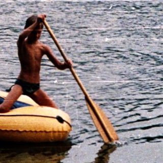 Two people rowing inflatable boat on calm lake.