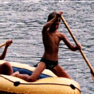 Two people rowing inflatable boat on calm lake.