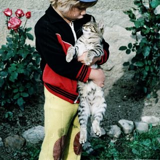 Child holding striped cat outdoors near plants.