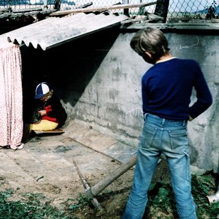 Child sitting under makeshift shelter, another child observing.