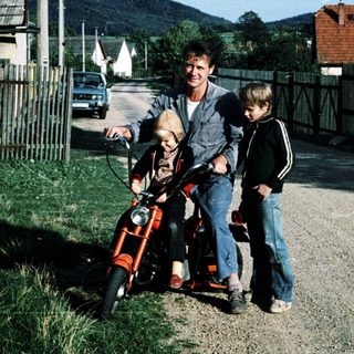 A man rides a red moped with two children beside him on a quiet road.