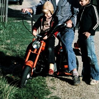 A man rides a red moped with two children beside him on a quiet road.