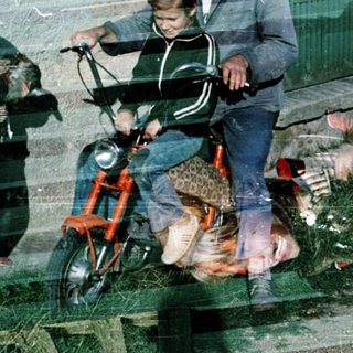 A man rides a moped with a child, another child stands nearby.