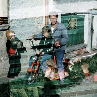 A man rides a moped with a child, another child stands nearby.