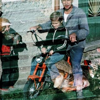 A man rides a moped with a child, another child stands nearby.