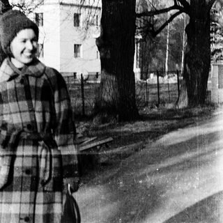 Old woman in plaid coat posing outdoors near bare trees.