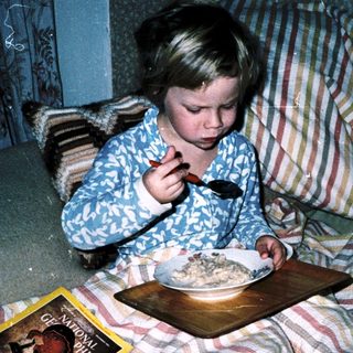 Child eating cereal in bed with book nearby.