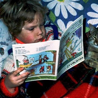 Child reading comic book with curious cat nearby.