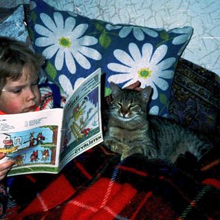 Child reading comic book with curious cat nearby.