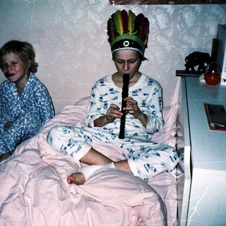 Two children in pajamas playing indoors with a feathered headdress.