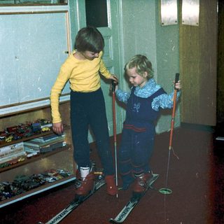 Two children indoors practicing skiing with poles.