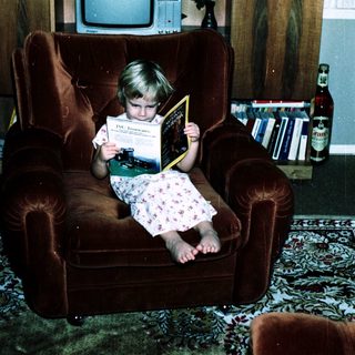 Child reading book on armchair in living room.
