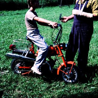 Young boy learning to ride a red lawn mower with adult supervision.