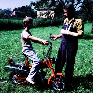 Young boy learning to ride a red lawn mower with adult supervision.