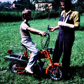 Young boy learning to ride a red lawn mower with adult supervision.