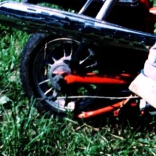 Young boy learning to ride a red lawn mower with adult supervision.