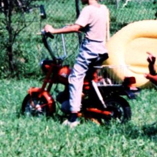 Child riding sidecar on moped through grassy field.