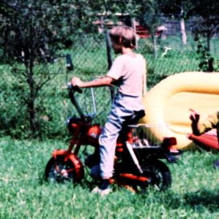 Child riding sidecar on moped through grassy field.