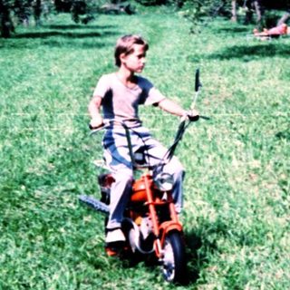 Child riding small lawn mower in grassy orchard.