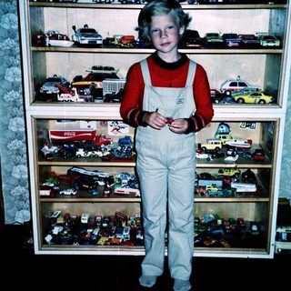 Boy in overalls poses with toy vehicles.