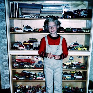 Boy in overalls poses with toy vehicles.