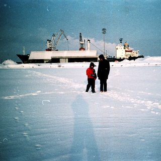 Two people standing on frozen sea near industrial ships.