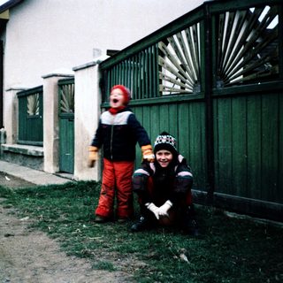 Two children dressed warmly posing outdoors near a fence.
