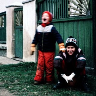 Two children dressed warmly posing outdoors near a fence.
