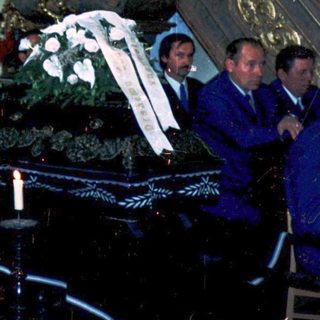 Funeral service with priest reading at open coffin draped in flowers.
