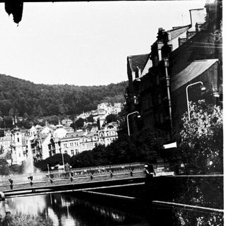 Historic town scene with canal, bridge, and hillside buildings.