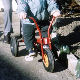 A woman assists a young child riding a red balance bike outdoors.