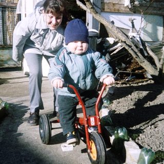 A woman assists a young child riding a red balance bike outdoors.