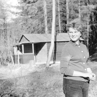 Woman standing outdoors near rustic cabin in forested area.