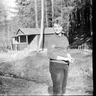 Woman standing outdoors near rustic cabin in forested area.