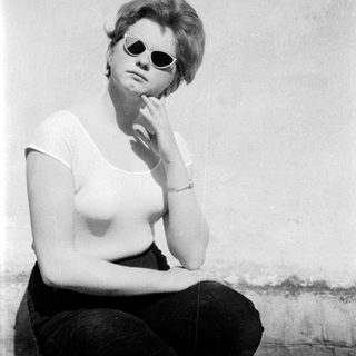 A woman in sunglasses sitting thoughtfully against a wall.