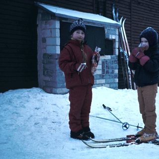 Two children holding skis by a snowy building.