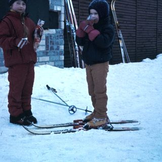 Two children holding skis by a snowy building.