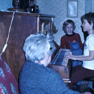 Family gathering with elderly woman playing piano, children and man listening.