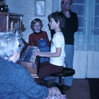 Family gathering with elderly woman playing piano, children and man listening.