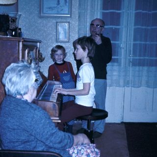 Family gathering with elderly woman playing piano, children and man listening.