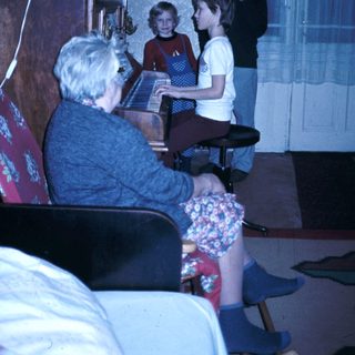 Family gathering with elderly woman playing piano, children and man listening.