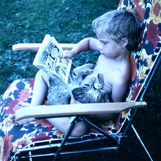 Shirtless child reading book with cat on chair.