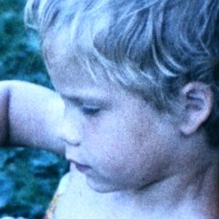 Shirtless child reading book with cat on chair.