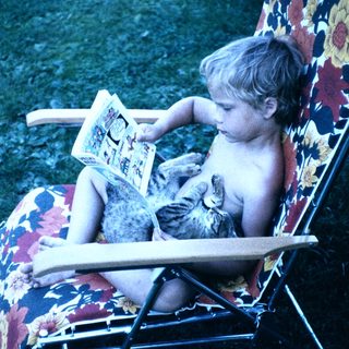 Shirtless child reading book with cat on chair.